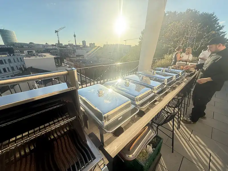 Catering-Aufbau mit Grill und Warmhaltebehältern auf einer Dachterrasse in der Sonne, daneben ein Koch