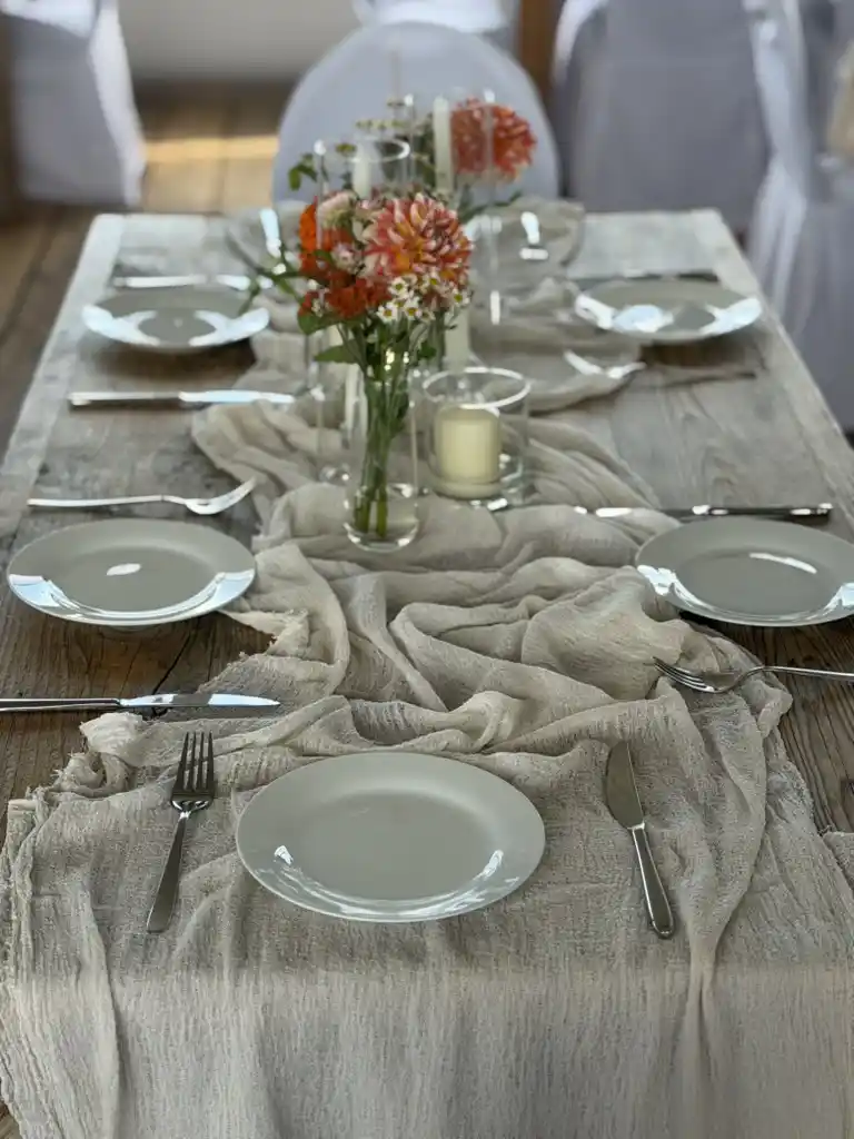 Gedeckte Hochzeitstafel mit Dahlienstrauß, Kerzenglas und Leinenläufer auf einem Holztisch